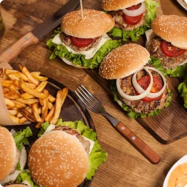 Several burgers with lettuce, tomato and onion beside a basket of fries on a wooden table.
