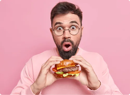 A surprised man in glasses holding a burger with both hands against a pink background