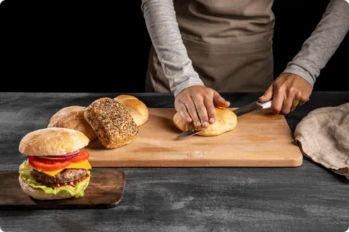 A cook in an apron slicing a brioche bun on a wooden board, with a finished burger beside it