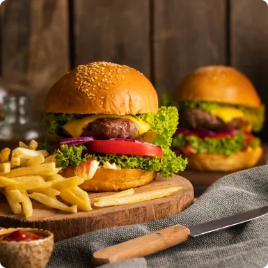 Two cheeseburgers on a wooden board with fries, a knife and a dish of ketchup.