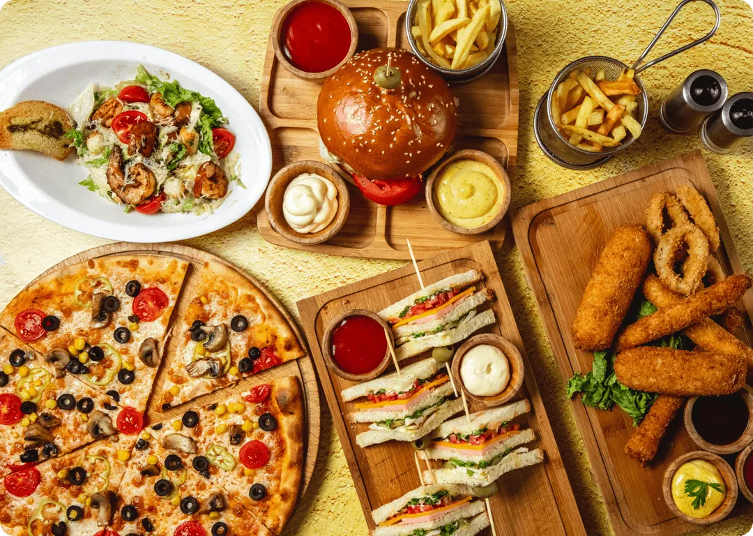 A table spread with a burger, pizza, sausages, sandwiches, fries and small bowls of sauce