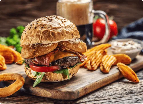 A burger on a toasted bun with onion rings, potato wedges and a dipping sauce on a wooden board