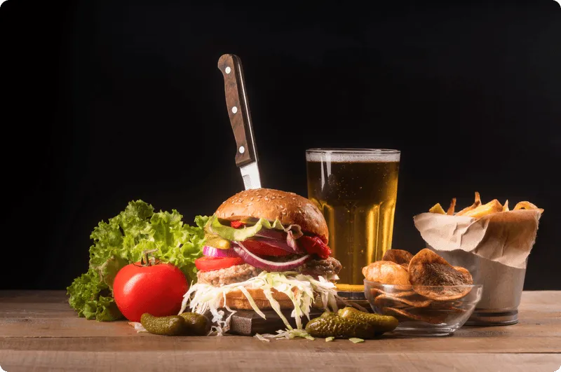 A tall burger held together with a steak knife, served with a glass of beer, fries and fresh vegetables