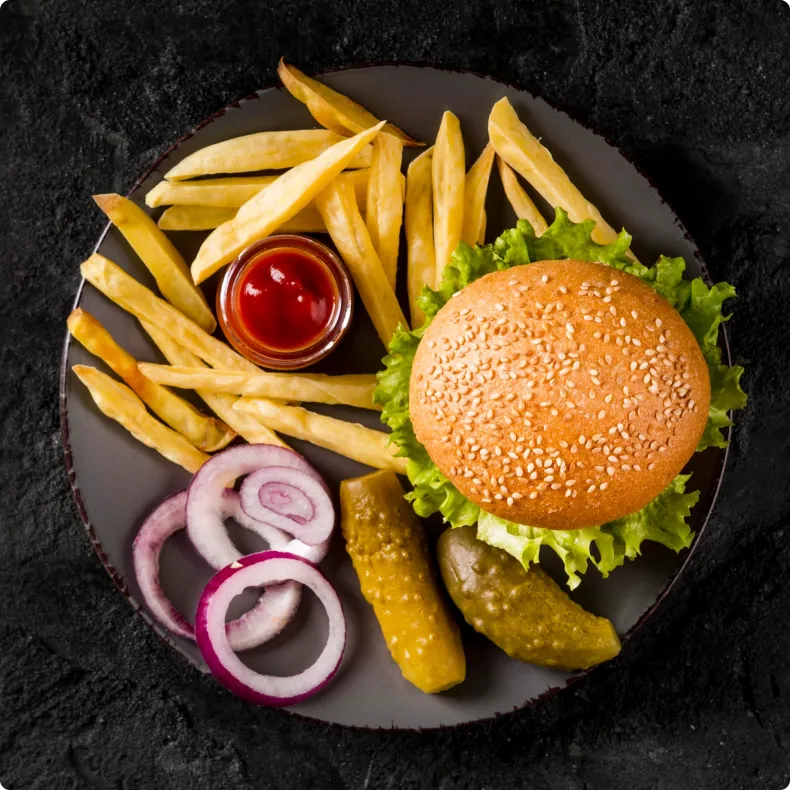 A burger on a plate seen from above, with fries, ketchup, pickles and red onion rings.