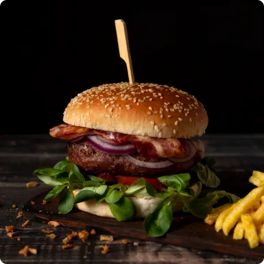 A beef burger with bacon, red onion and greens, pinned with a wooden skewer, with fries on the side.