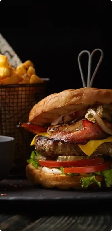 A tall cheeseburger with bacon and tomato in front of a basket of fries.