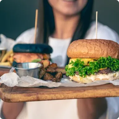 A server holding a wooden board with two burgers and a dish of potato wedges.