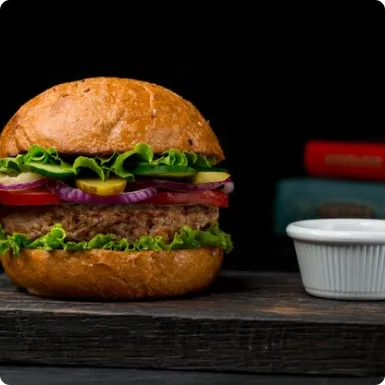 A burger with lettuce, tomato, red onion and pickles on a wooden board, with a dish of sauce.