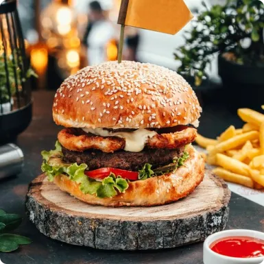 A beef patty topped with crispy chicken and sauce on a wooden slice, with fries and ketchup behind it.