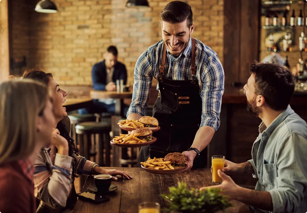 A smiling server bringing plates of burgers and fries to a table of friends in a brick-walled dining room